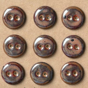 May include: A set of nine round, two-hole buttons featuring a glossy, variegated glaze in shades of deep brown, copper, and dark charcoal. Each button has a smooth, reflective surface with subtle color variations and a distinct, circular shape.