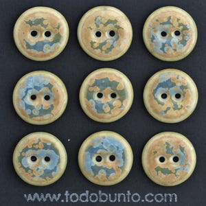 Può includere: Nove bottoni rotondi in ceramica, ciascuno con due fori, disposti in una griglia 3x3. I bottoni hanno una base color crema e un design screziato nei toni del blu, arancione e verde. Il testo "todobunto.com" è in basso.