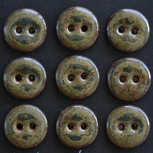 Puede incluir: Un conjunto de nueve botones de cerámica redondos dispuestos en una cuadrícula de tres por tres. Cada botón presenta un diseño de dos agujeros y un acabado de esmalte moteado en tonos verde oliva, marrón oscuro y bronceado con textura brillante.