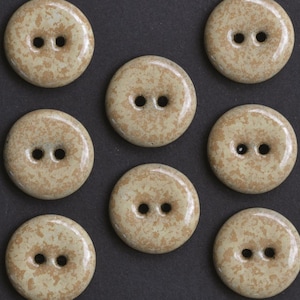 May include: Seven round buttons with a smooth, glossy finish. Each button features a light beige base colour with a mottled, tan-coloured speckled pattern and two centred holes for attachment. The buttons are arranged in a grid-like formation.