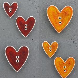 Puede incluir: Un conjunto de seis botones en forma de corazón con un acabado de esmalte agrietado. Tres botones son de color rojo oscuro y tres son de color amarillo anaranjado vibrante. Cada botón tiene dos agujeros verticales y un borde blanco.