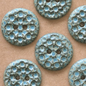 May include: A collection of round, two-hole buttons featuring a textured, raised floral pattern. The buttons have a glazed finish with a color palette of light blue and muted grey-green, creating a mottled appearance across the surface.