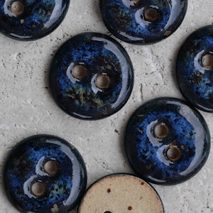 May include: A collection of round, two-hole buttons featuring a glossy, dark blue and black glaze with mottled, crystalline patterns. Each button has a smooth, reflective surface and a visible, unglazed tan ceramic base on the underside.