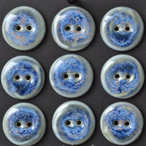 May include: A set of nine round, two-hole buttons arranged in a grid. Each button features a glossy, glazed finish with a swirling pattern of deep blue, light blue, and cream, accented by small, speckled copper-colored dots across the surface.