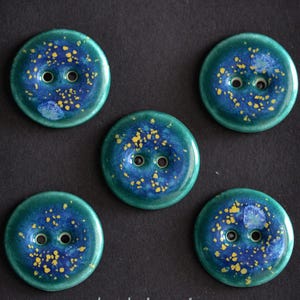 May include: Five round buttons featuring a glossy finish with a deep blue and teal gradient base. Each button is decorated with scattered yellow speckles and small white accents, and contains two central holes for attachment to garments.