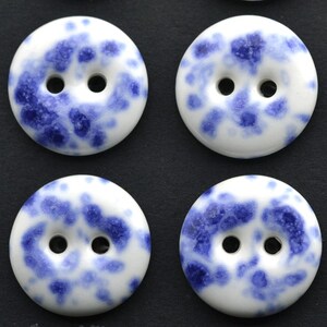 May include: Four round white buttons with a blue speckled pattern. Each button has two holes for sewing. The buttons are arranged in a square formation against a dark background.