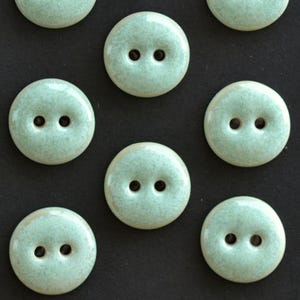 Puede incluir: Una colección de botones redondos de dos agujeros con un acabado liso y brillante en color verde menta pálido, con sutiles motas blancas por toda la superficie de cada botón individual, dispuestos en un patrón que se repite.