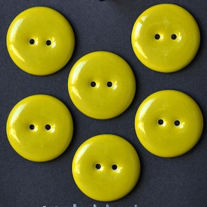 May include: Six vibrant yellow, circular buttons, each with two holes, displayed on a dark grey background. The buttons have a glossy sheen, and the holes are symmetrically placed. Perfect for sewing and craft activities.