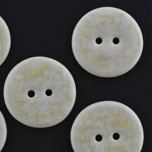 May include: A collection of round, two-hole buttons featuring a mottled, off-white and pale yellow surface texture. Each button has a smooth, circular edge and two centered holes for attachment, arranged in a scattered pattern across the frame.