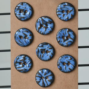 Puede incluir: Un conjunto de ocho botones redondos de cerámica dispuestos sobre una tarjeta marrón. Cada botón presenta un esmalte azul brillante con patrones de color marrón oscuro en forma de ramas en la superficie. Cada botón tiene dos agujeros.