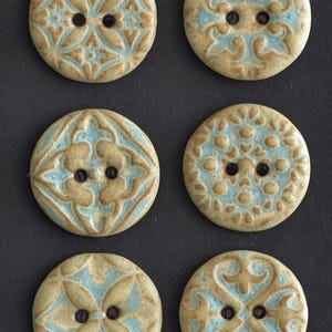 May include: Six round ceramic buttons with intricate, raised patterns in light blue and tan. Each button has two holes for sewing. The buttons feature various floral and geometric designs, suitable for sewing projects.
