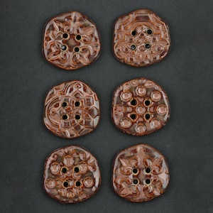 Op de afbeelding: Zes bruine keramische knopen met ingewikkelde reliëfdesigns. Elke knoop heeft een uniek patroon, waaronder bloemen- en geometrische motieven, en heeft vier of zes gaten om op te naaien. De knopen hebben een glanzende afwerking en zijn geplaatst op een donkere achtergrond.