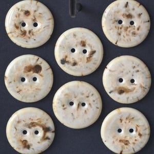May include: A collection of eight round buttons with two holes each. The buttons have a creamy white base color with brown marbling. The buttons are arranged on a dark background.