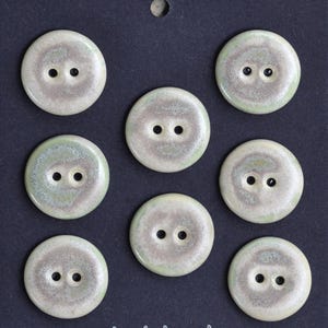 Könnte beinhalten: Acht runde Keramikknöpfe mit jeweils zwei Löchern, auf dunklem blauem Hintergrund. Die Knöpfe haben eine gesprenkelte, cremefarbene und hellgrüne Glasur. Jeder Knopf hat einen Durchmesser von etwa 2,5 cm.
