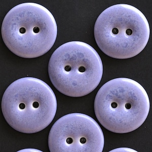 May include: A collection of round, two-hole buttons in a light lavender color with a subtle, mottled, speckled texture across the surface. Each button features a smooth, slightly domed shape with two centered holes for attachment to fabric.