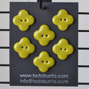 May include: Six yellow, four-petal shaped buttons with two holes each are displayed on a dark gray card. The buttons have a glossy finish. The card has the website address todobunto.com and an email address printed on it.