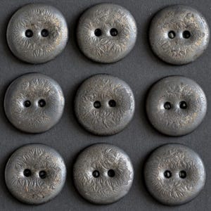 May include: A set of nine circular, dark gray buttons arranged in a three-by-three grid. Each button features a two-hole center design and a textured, radiating pattern across the surface, resembling fine, etched lines or organic, fibrous details.