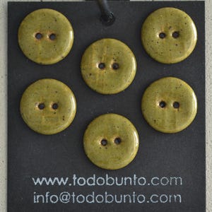 Puede incluir: Cinco botones redondos de cerámica de color verde oliva con un acabado brillante y moteado, cada uno con dos agujeros centrales. Los botones están dispuestos sobre una tarjeta de presentación negra con el texto www.todobunto.com y info@todobunto.com.