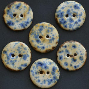 May include: Six ceramic buttons with a speckled design. The buttons are round, with a cream-colored base and blue speckles. Each button has two holes for sewing and a dark brown edge. The buttons are arranged on a dark gray surface.