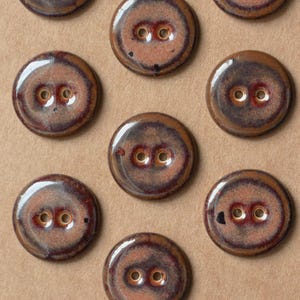 May include: A selection of ceramic buttons, each with a glossy finish and two holes. The buttons are circular and feature a brown and reddish-brown colour palette, with unique patterns created by the glaze. These buttons are suitable for sewing and craft projects.