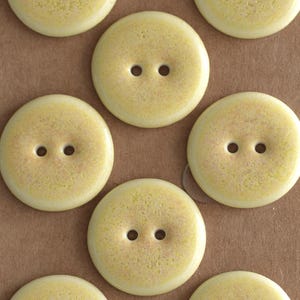 May include: A collection of round, two-hole buttons featuring a pale yellow color with a subtle, speckled, and textured surface finish. Each button has a smooth, slightly raised edge and a flat center, arranged in a repeating pattern across the frame.