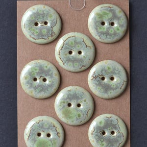 May include: A collection of eight round, ceramic buttons. Each button is light green with darker green and brown mottled patterns. The buttons have two holes for sewing and are arranged on a brown card.