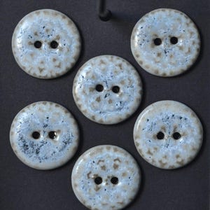 May include: Six round, light blue buttons with two holes each. The buttons have a speckled, textured design in shades of brown and white around the edges. The buttons are arranged on a dark gray surface.