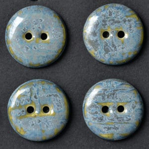 Puede incluir: Un conjunto de cuatro botones redondos de dos agujeros con un esmalte brillante y moteado en tonos azul, gris y amarillo. Cada botón tiene una superficie circular lisa con un patrón irregular distintivo del acabado esmaltado.