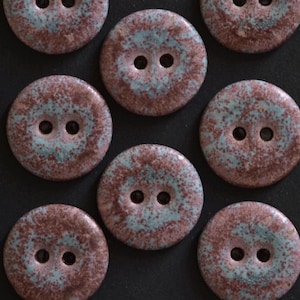 May include: A collection of seven round, two-hole buttons featuring a speckled glaze finish. Each button displays a mottled pattern of earthy brown and soft teal tones, creating a textured appearance across the circular surfaces.