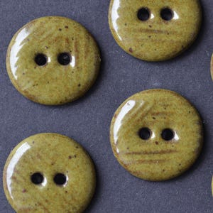 May include: Close-up of several round, olive-green buttons with two holes each. The buttons have a glossy finish and a speckled, textured appearance. These decorative buttons are ideal for sewing and craft projects.
