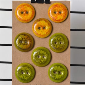 May include: A collection of eight round buttons in shades of yellow and green, each with two holes for sewing. The buttons have a textured, marbled appearance, with variations in color and pattern. They are arranged on a brown card.