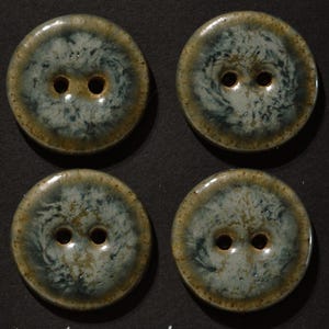 May include: Four circular buttons arranged in a two-by-two grid. Each button features a glossy, mottled glaze with a blend of sage green, dark blue, and earthy brown tones. Every button has two central holes and a smooth, rounded edge profile.