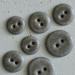 May include: A collection of seven round, dark gray buttons with a speckled, textured finish. Each button features two central holes for attachment. The buttons vary in size, displaying a smooth, slightly glossy surface with subtle brown undertones.