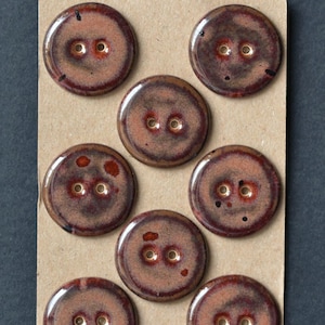 May include: A collection of eight round, glazed buttons. The buttons are a mottled brown with dark brown edges and two holes. They are arranged on a tan card.