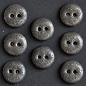 May include: A set of seven circular, metallic-toned buttons arranged in a grid pattern. Each button features a textured, uneven surface with a dark grey finish and two central holes for attachment, providing a consistent look across all pieces.