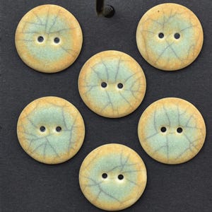 May include: Six round buttons featuring a two-hole design. Each button has a light blue center with a dark blue crackle pattern and a tan border. The buttons are arranged in a circular pattern against a dark gray background.