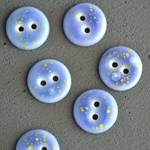 May include: Six round buttons featuring a smooth light blue surface with scattered yellow speckles. Each button has two central holes for attachment. The buttons have a glossy finish and a uniform circular shape with a slightly raised center.