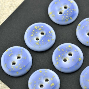 May include: A collection of round, two-hole buttons featuring a smooth, light blue surface with scattered yellow speckles. Each button has a glossy finish and a slightly raised center, arranged in a pattern on a dark, textured surface.