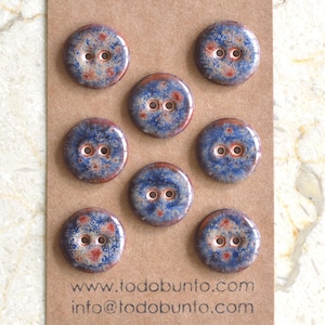 May include: A set of eight ceramic buttons with a glossy finish. The buttons are round, with a blue base color and speckled red and white accents. Each button has two holes for sewing. The buttons are displayed on a brown card with the website and email address.