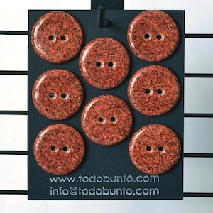 Op de afbeelding: Negen ronde, gespikkelde, oranjerode knopen met elk twee gaten worden getoond op een zwarte kaart. De knopen hebben een glanzende afwerking. De kaart bevat het websiteadres www.todobunto.com en een e-mailadres.