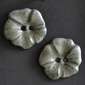 May include: Two flower-shaped buttons with a mottled grey and white finish. Each button features a five-petal design with a slightly raised center and two central holes for attachment. The surface has a textured, weathered appearance.