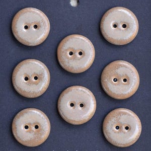 May include: A collection of eight round, two-hole buttons. The buttons have a light beige center with a darker, mottled brown edge. They are arranged on a dark blue surface.
