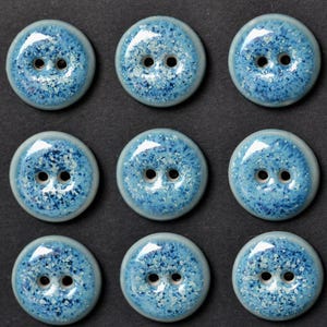 May include: Nine round, light blue ceramic buttons with two holes each. The buttons have a speckled design with white and darker blue accents. The buttons are arranged in a three-by-three grid on a dark background.