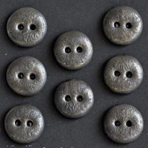 Puede incluir: Primer plano de ocho botones antiguos de color plateado. Cada botón es redondo, con dos agujeros y una superficie texturizada. Los botones están dispuestos sobre un fondo oscuro, mostrando su brillo metálico y detalles intrincados.