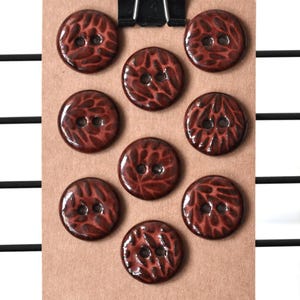 May include: A collection of nine round, dark red buttons with a textured, leaf-like pattern. Each button has two holes for sewing. The buttons are arranged on a brown card, held by a black clip.