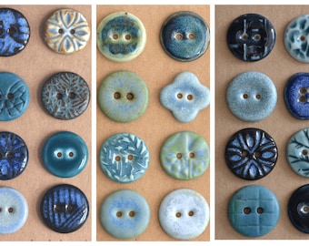May include: A collection of ceramic buttons in various shades of blue, teal, and cream. The buttons feature diverse designs, including floral patterns, textured surfaces, and unique shapes. Each button has two holes for sewing.