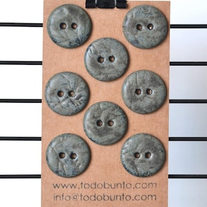 May include: Seven gray, round buttons with two holes each are arranged on a brown card. The buttons have a textured surface and a leaf-like design. The card is held by a black clip. The website address www.todobunto.com and email info@todobunto.com are printed at the bottom.