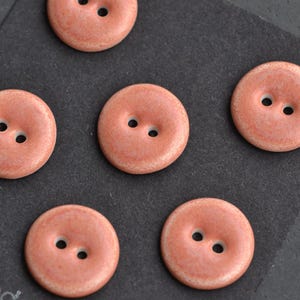 May include: Close-up of six round, coral-colored buttons with two holes each. The buttons are arranged on a dark gray surface. The buttons have a slightly textured appearance.