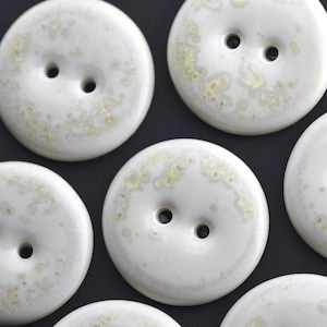 May include: Close-up of several white ceramic buttons with two holes each. The buttons have a speckled, golden-yellow pattern on the surface. The buttons are round and have a glossy finish, set against a dark background.