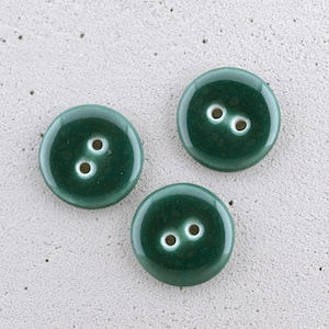May include: Three round, dark forest green buttons, each featuring a smooth, glossy finish and two central holes for attachment. The buttons have a slightly mottled colour variation within the green surface, creating a consistent appearance.